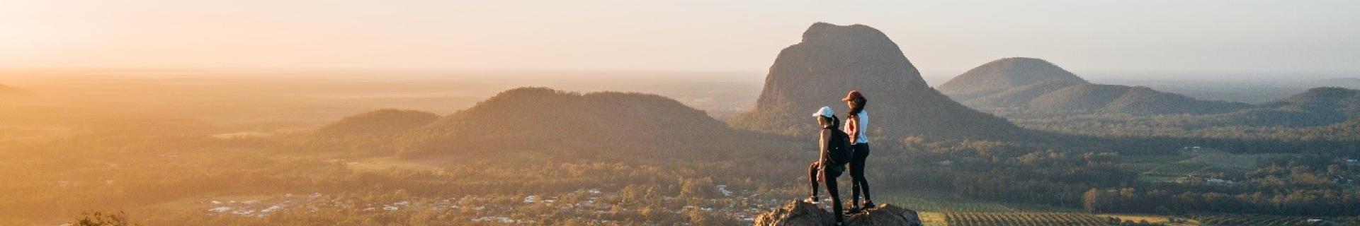 Two hikers stand on a hill-top looking into the distance. 