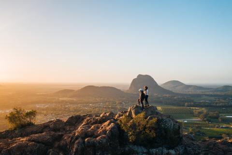 Two hikers stand on a hill-top looking into the distance. 