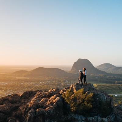 Two hikers stand on a hill-top looking into the distance. 