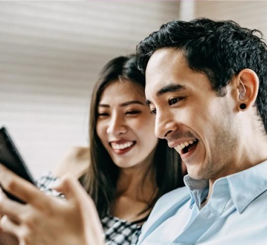 A hearing impaired man is sitting on a couch with a woman. They are both smiling and watching the screen of a smart phone while entering text.