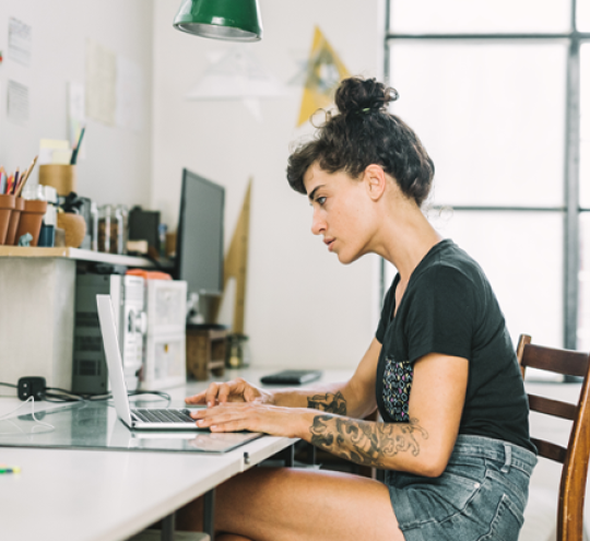 A woman sits at a work bench in a well-lit room that also includes a sink, cupboards and many green houseplants. The bench she sits at is crowded with a large, glossy open book; an open exercise book covered with notes and designs; artists’ pencils; etc.