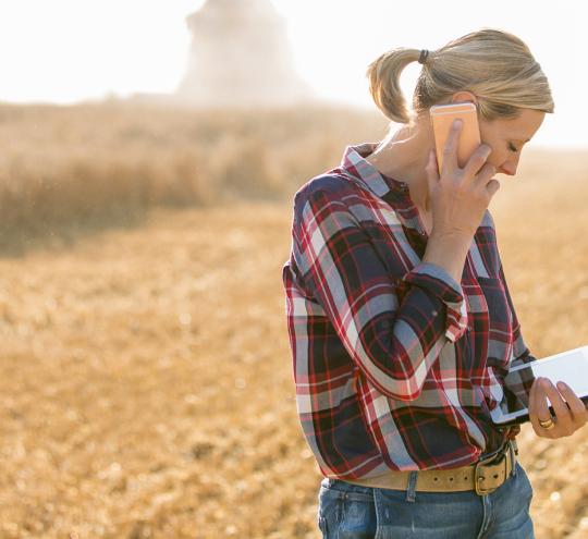 Woman farmer in a field talking on a mobile phone and looking at a mobile device in her left hand.