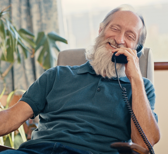 An older-aged man on a landline telephone is enjoying a casual conversation in his home.