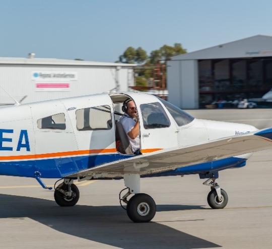 A small plane on the ground, featuring a pilot in the front seat with the door half open. Sheds are in the background, with clear skies.