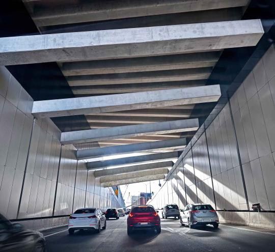 A range of different cars driving along a three lane road. The cars appear to be exiting an underpass or tunnel which has large metal beams formed overhead as an artistic structure. 