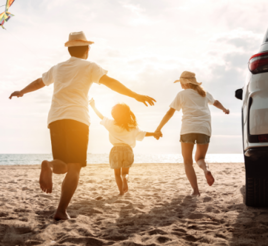 A family holiday hands and running on a beach with a kite and a car.