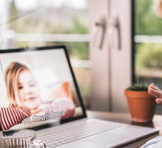 The screen of a lap top shows a young girl with the caption “Hi Grandma”. An elderly woman waves at the screen.