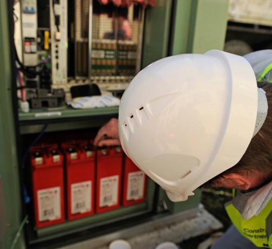 Person wearing a hard hat inspecting shelves in a cupboard with telecommunication tools.