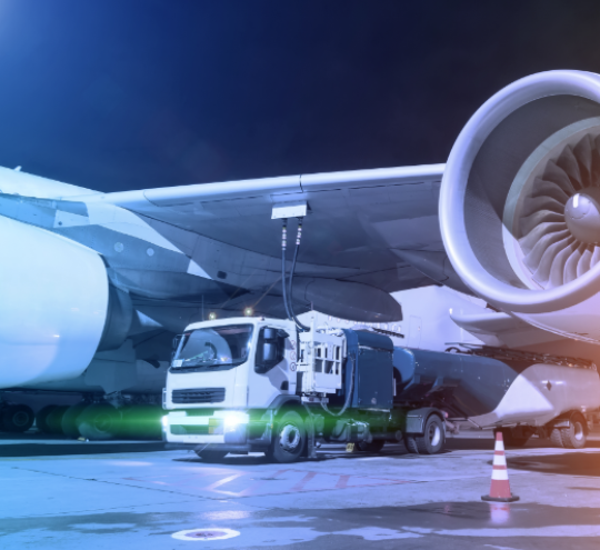 a plane being refuelled at night through the underside of its wing by a fuel truck with green headlights