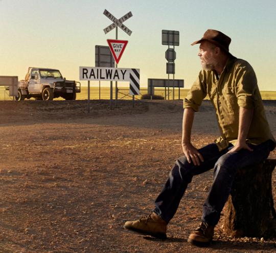 A man in an Akubra hat sits beside a regional road with railway crossing signage and a ute stopped near the crossing.