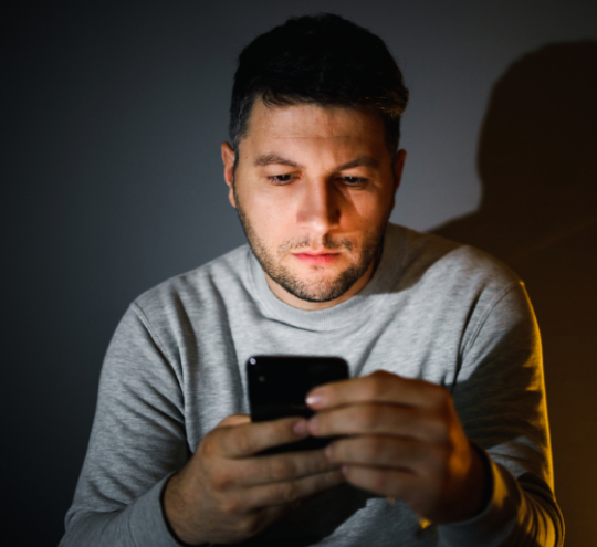 Man sitting in a dark room looking at his smartphone.