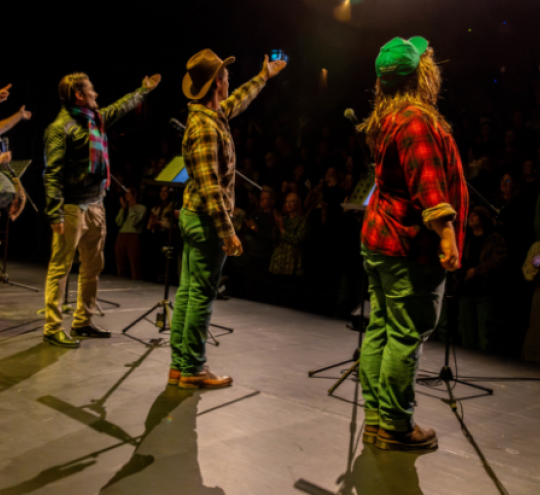 Six actors stand on a lit stage facing a seated crowd