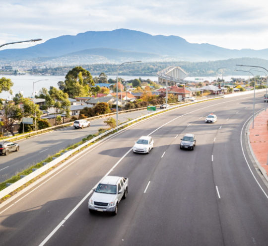 Overhead view of vehicles driving on a four-lane road in both directions with trees and mountains in the distance. 