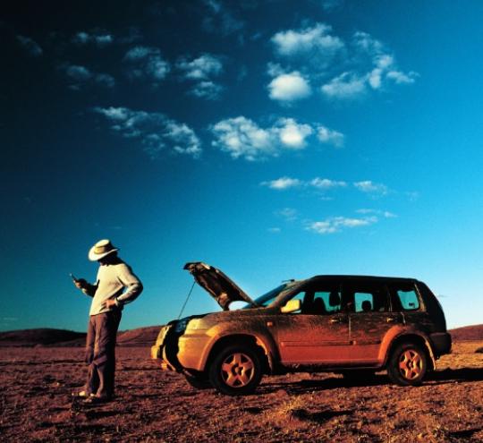 Man with a mobile phone in his hand standing next to his broken down car in the outback. 