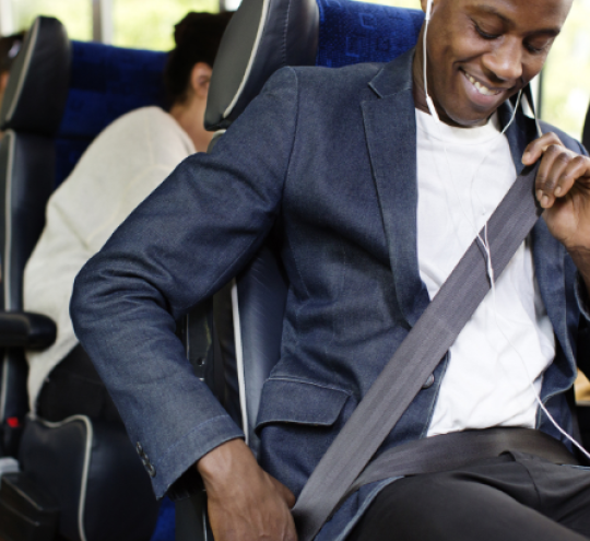 A man in a blue jacket and white t-shirt fastens his seatbelt on a bus, with other passengers in the background.