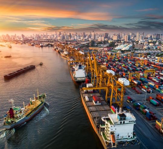 Ships in a body of water and docked beside an array of shipping freight containers, with a city skyline in the background