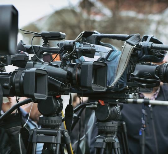 Several news cameras on tripods at an outdoor press conference.