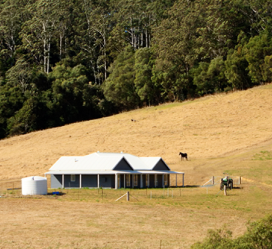 Rural house on the side of a hill in an open area below a forested area. The grass is dry.