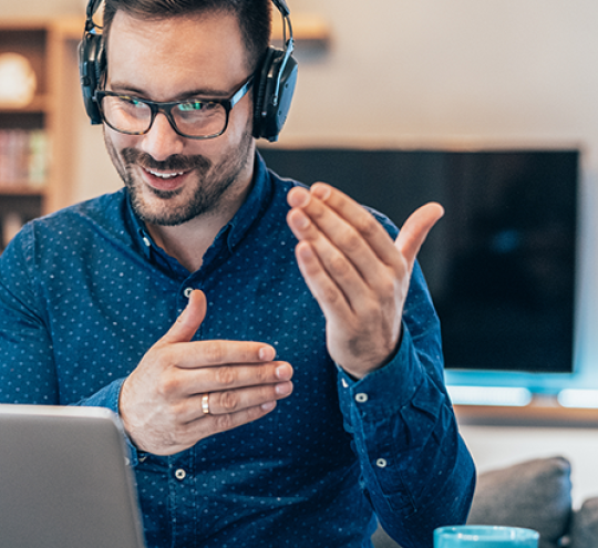 Man sitting at a desk. He is wearing headphones and looking at a laptop. It looks like he's talking to the laptop.