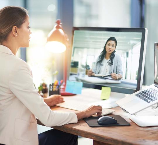 Business woman sitting at desk talking with another woman on the computer screen.