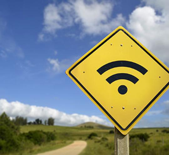 Wifi symbol on a yellow diamond shaped road sign in a rural setting next to a dirt road