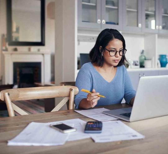 Woman sitting in front of a laptop with papers and a mobile device and calculator beside her. She is in her home.