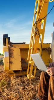 Farmer counts yields on a laptop whilst sitting in the tyre of a tractor in the middle of a paddock with another blue tractor in the background.