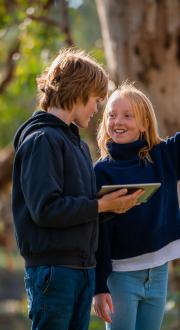 A young boy and girl standing outside in a park holding a digital tablet. 