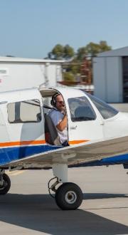 A small plane on the ground, featuring a pilot in the front seat with the door half open. Sheds are in the background, with clear skies.