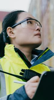 Asian female worker with clipboard and safety glasses looking up at a shipping containers stacked.