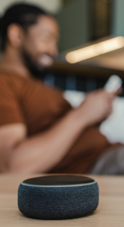 In the background a man sits on the couch, in the forefront a smartspeaker sits on a coffee table.