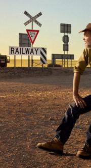 A man in an Akubra hat sits beside a regional road with railway crossing signage and a ute stopped near the crossing.