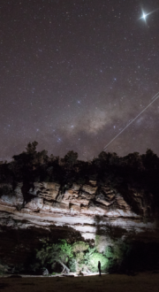 Campfire at cliffside looking up at a starry night and a low earth orbit satellite star train. 