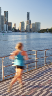 A woman jogs along a riverside footpath with colourful art sculptures in the foreground. In the background are established trees, and a city skyline across the river.