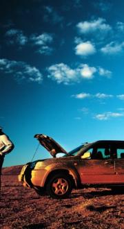 Man with a mobile phone in his hand standing next to his broken down car in the outback. 