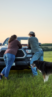 Couple leaning against the hood of their car looking at a laptop in a green field