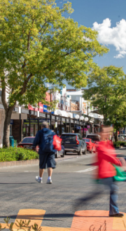 Four people walking on a street in a regional town setting, featuring older-style buildings with store-fronts, cars parked out the front of the stores, and trees along the sides of the street. 