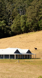 Rural house on the side of a hill in an open area below a forested area. The grass is dry.