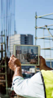 A worker in high vis jacket and hard hat holds up a tablet in front of them looking at at construction site