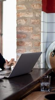 Woman drinking coffee at cafe bar with laptop in front of her. Man behind the cafe counter serving her.