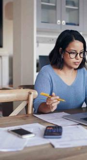 Woman sitting in front of a laptop with papers and a mobile device and calculator beside her. She is in her home.