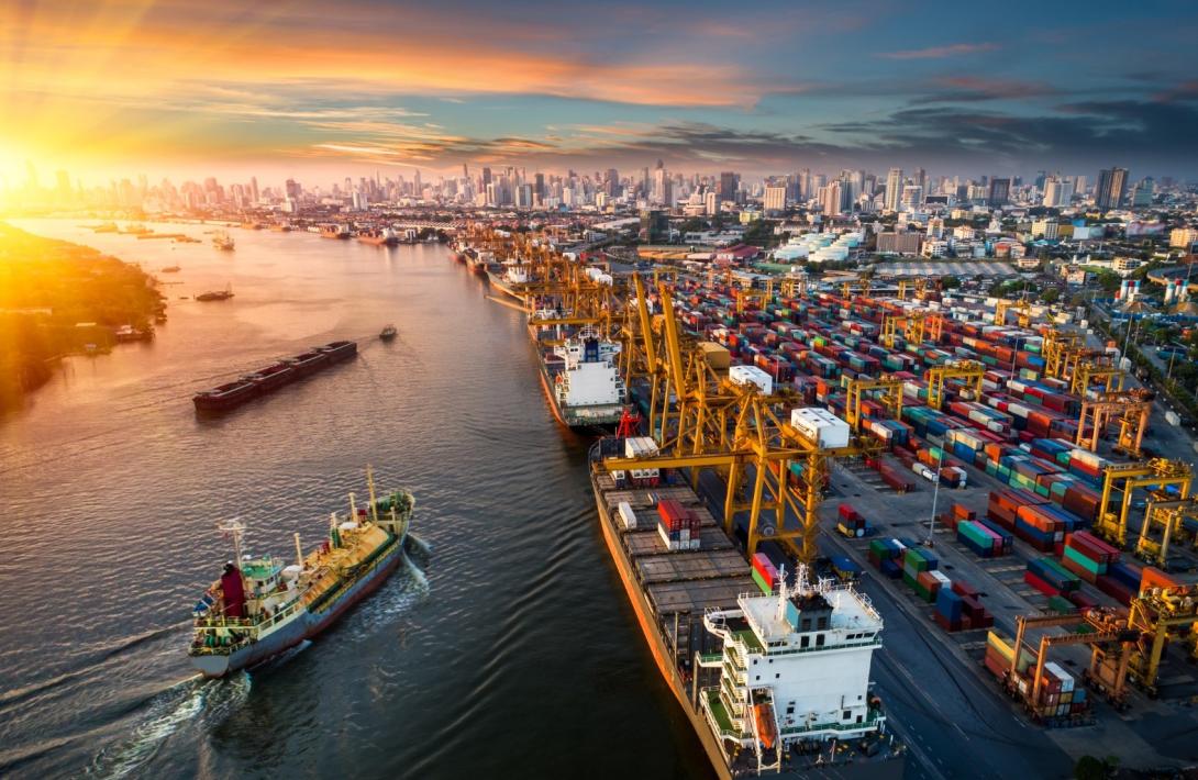 Ships in a body of water and docked beside an array of shipping freight containers, with a city skyline in the background